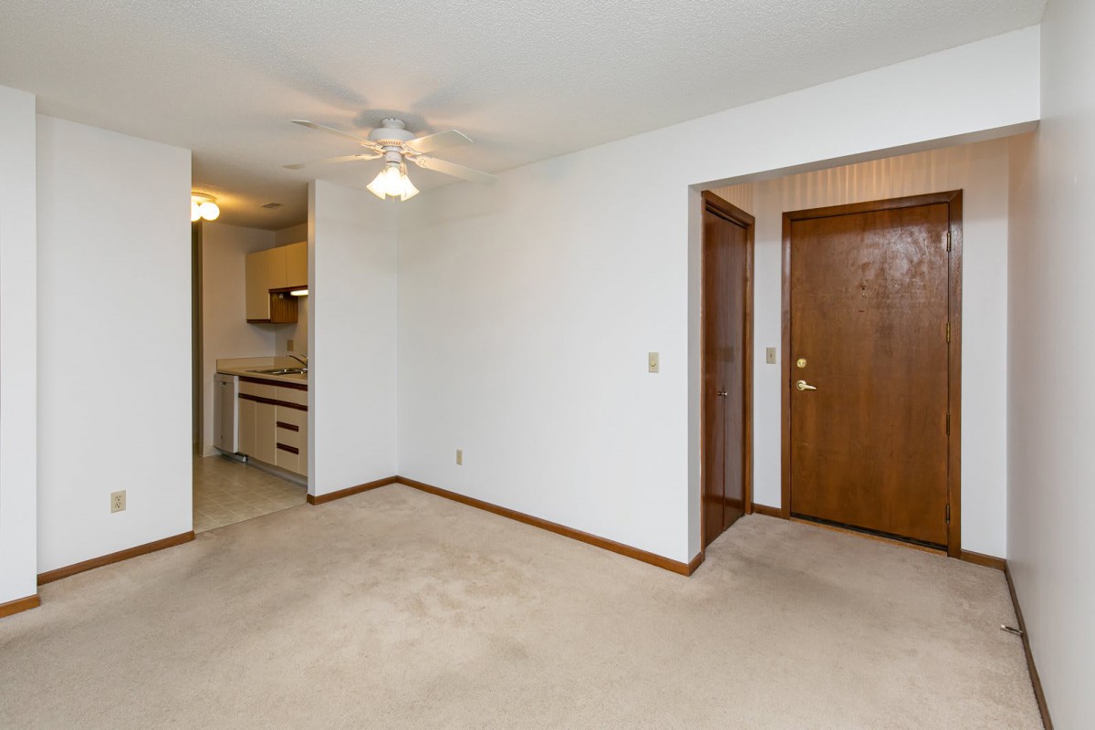 A simple apartment interior featuring a living room with carpet flooring, white walls, a ceiling fan, and a view of the kitchen and entrance door.