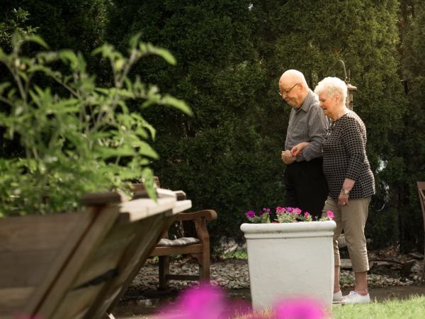 An elderly couple enjoying a stroll in the garden, surrounded by lush greenery and potted plants.