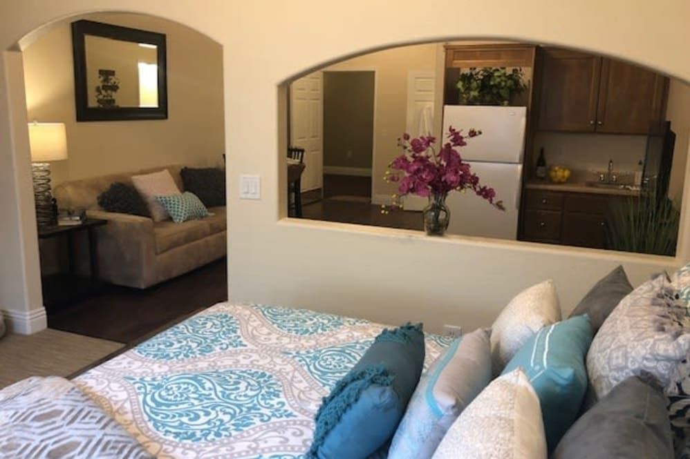 A cozy open-plan living space featuring a bedroom with a patterned quilt and colorful pillows, connected to a living area with a sofa and floral arrangement.