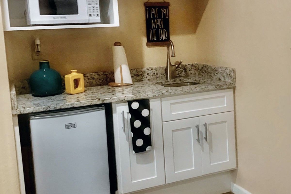 A small kitchen area featuring a granite countertop, white cabinets, a sink, microwave, and fridge.