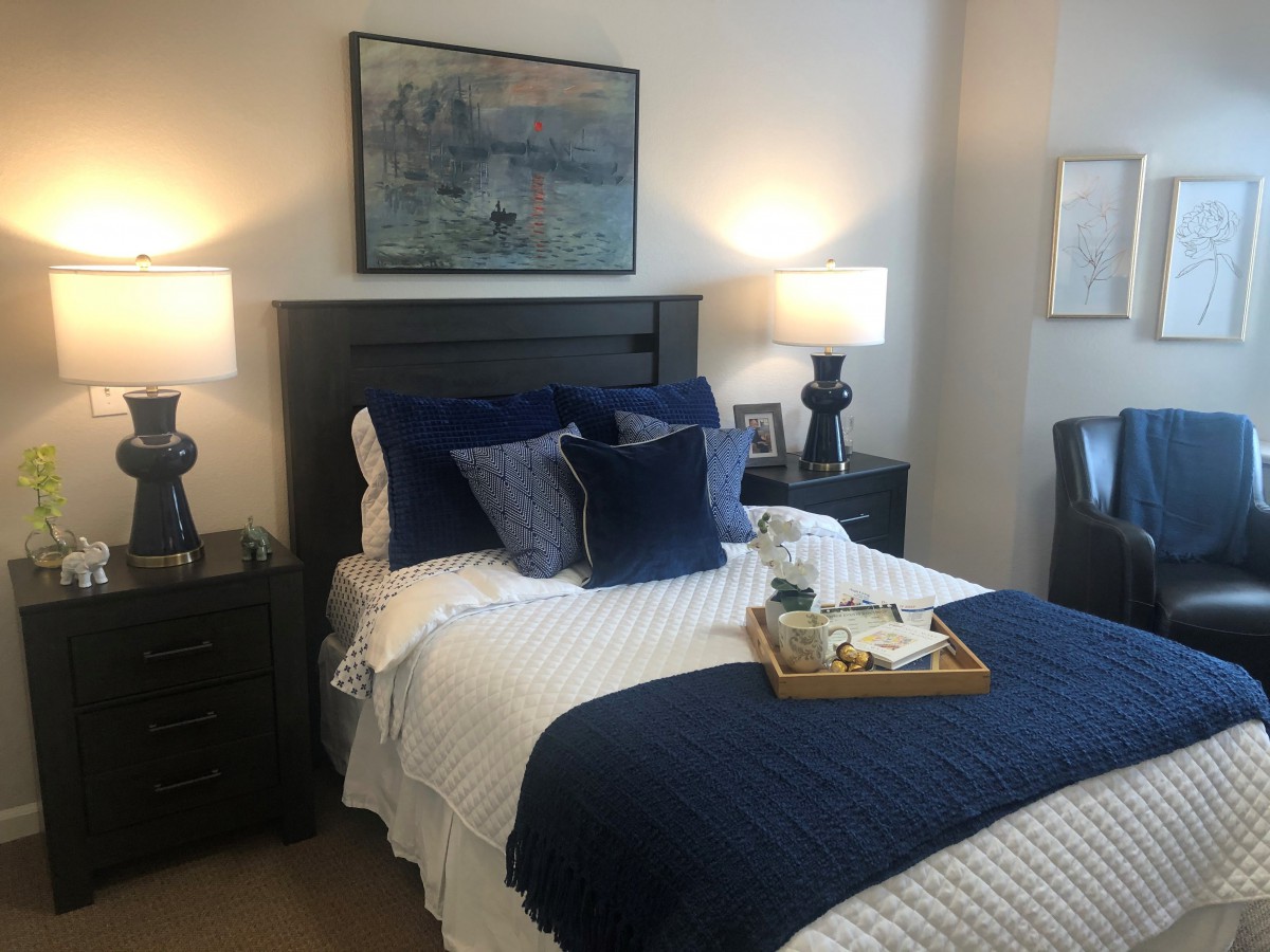 A cozy and well-decorated bedroom featuring a neatly made bed with blue and white bedding, accented by two bedside lamps and artwork above the headboard.