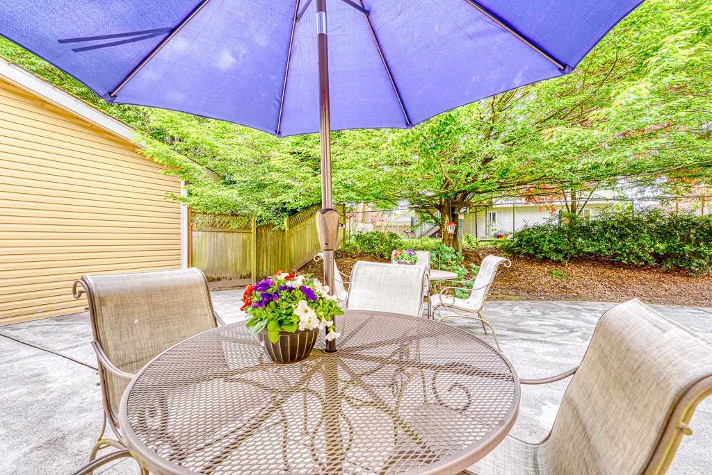 A cozy patio setup with a table, chairs, and a large umbrella surrounded by greenery.