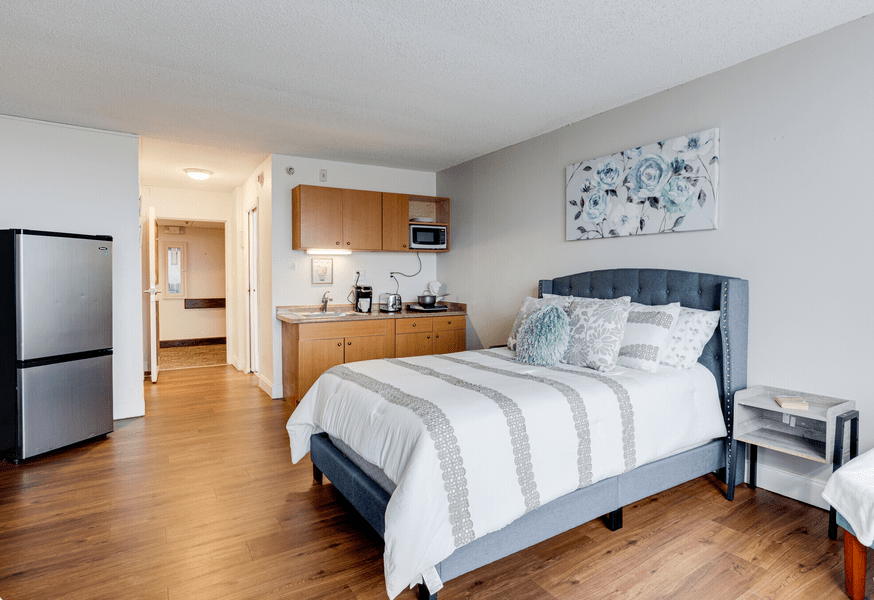 A modern bedroom with a neatly made bed featuring striped bedding, adjacent to a small kitchenette in an open-plan apartment.