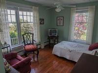 A cozy bedroom with large windows, featuring a bed, chairs, and a television on hardwood flooring.