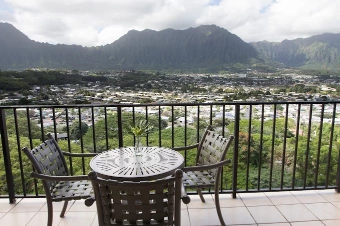 A scenic view from a balcony overlooking mountains and a valley with a table and chairs in the foreground.