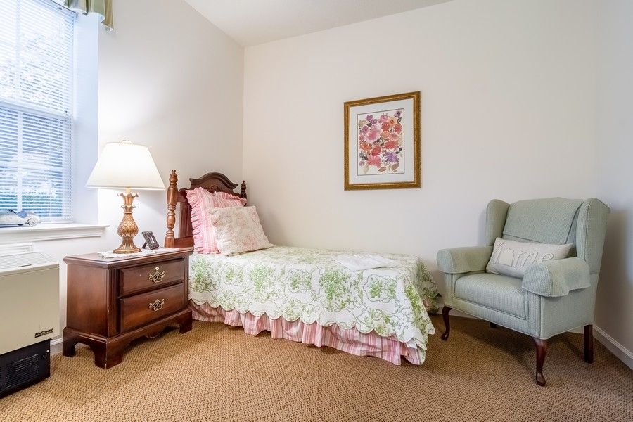 A cozy bedroom with a single bed, featuring floral bedding and a nightstand with a lamp. An armchair is placed nearby under decorative wall art.