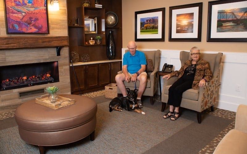 An elderly couple is seated in a cozy living room with their dog resting on the floor.