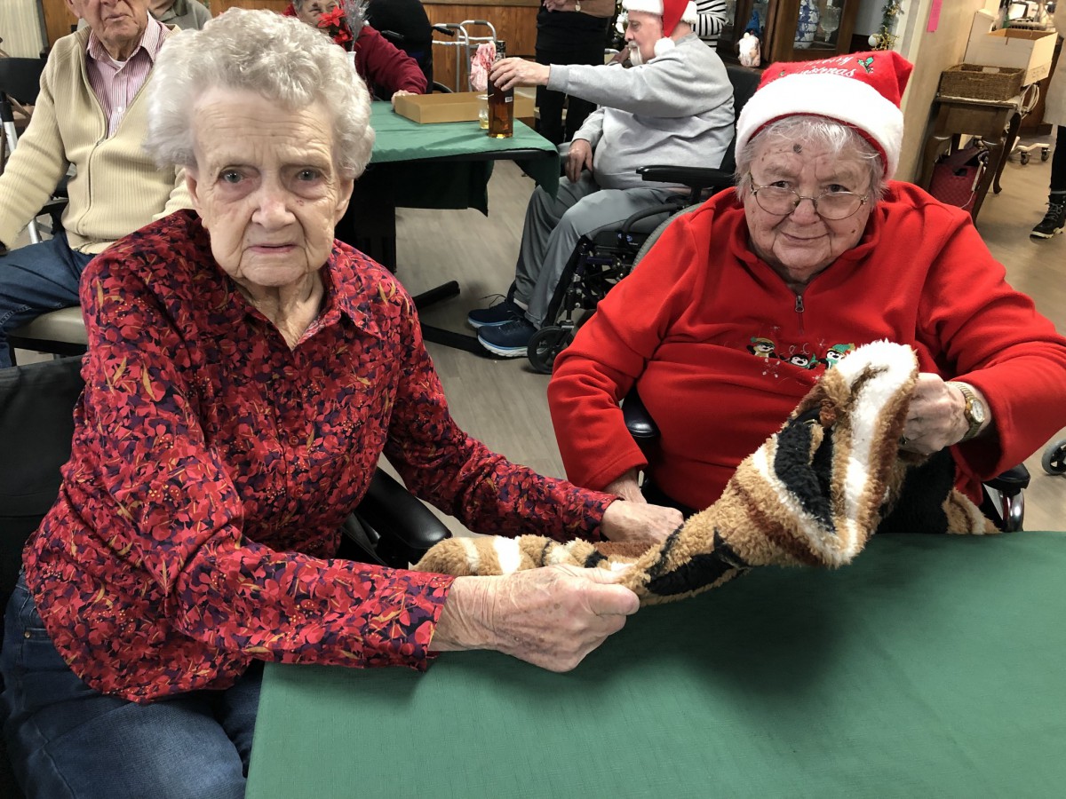 Two elderly people celebrate the holidays indoors, with one wearing a Santa hat and both holding a snake.
