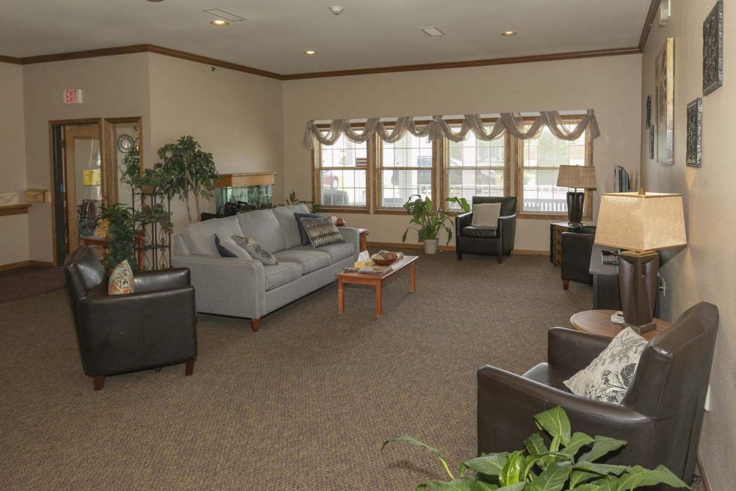 A cozy living room with sofas, chairs, and a coffee table surrounded by large windows and indoor plants.