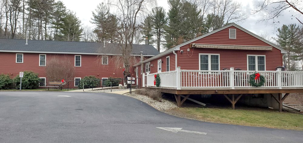 A red house with a white porch situated along a paved road, surrounded by trees.
