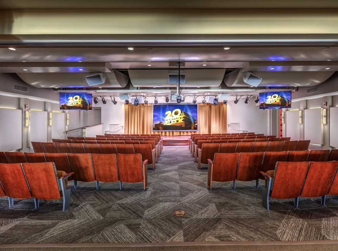 A spacious auditorium with red seating and a stage, equipped with television screens for presentations.