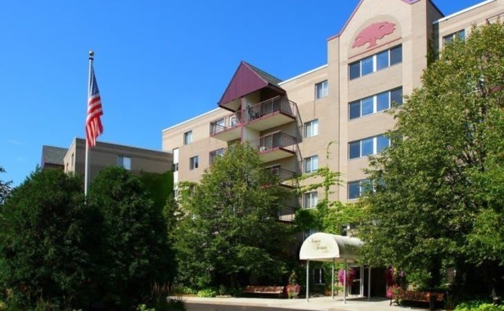 A multistory residential apartment building with balconies and surrounding trees, featuring an American flag at the entrance.