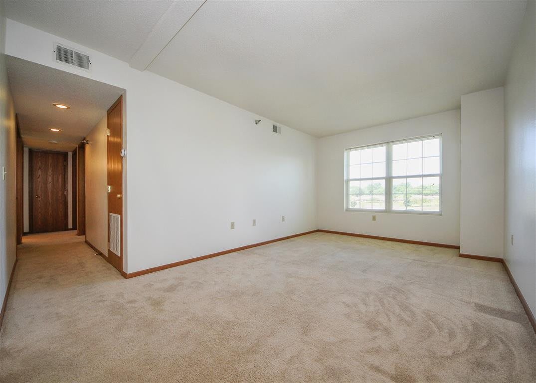 An empty room with white walls and a carpeted floor, featuring a large window letting in natural light.
