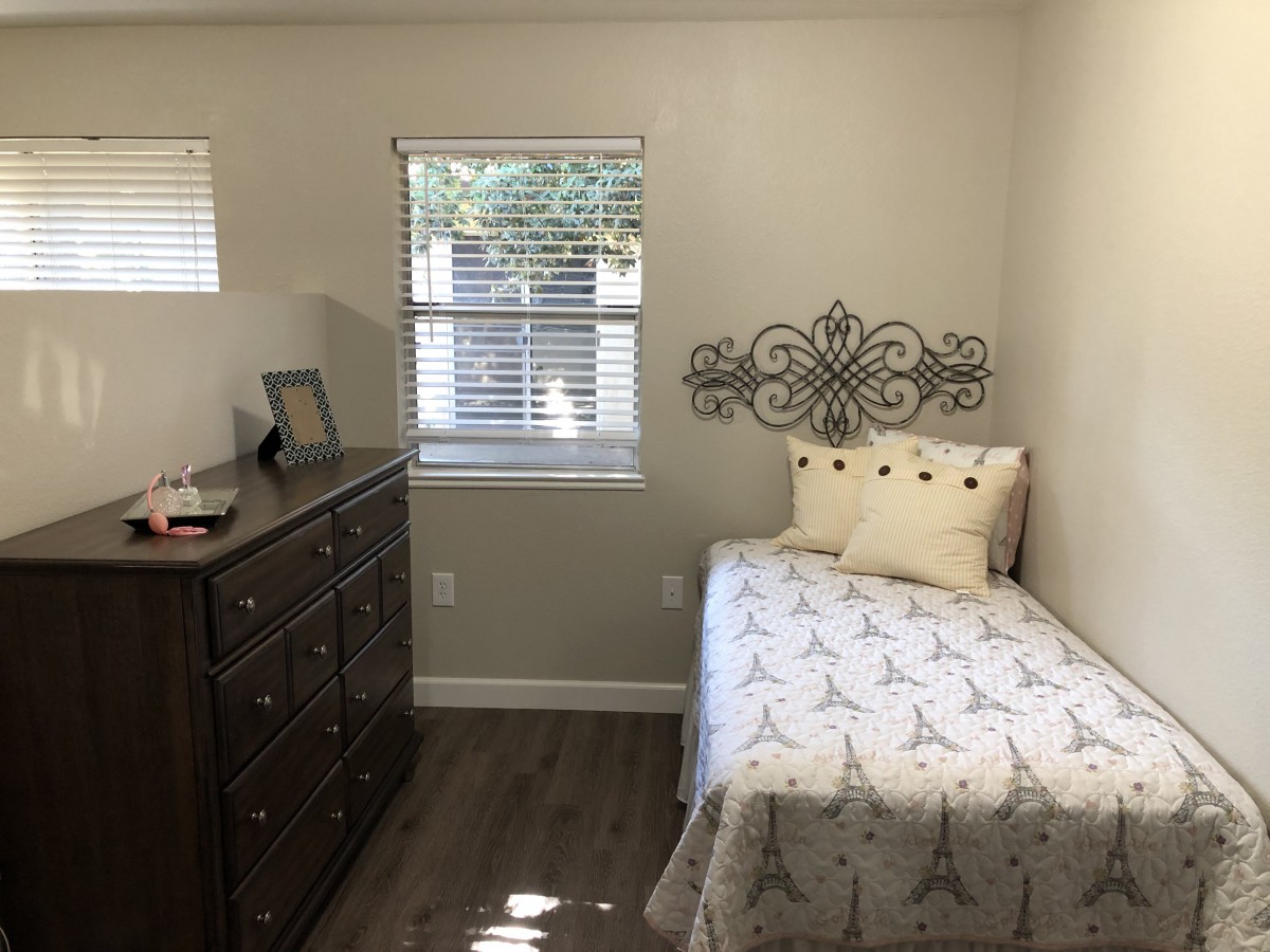 A cozy, minimalistic bedroom with a single bed and dresser, bathed in natural light from the window.