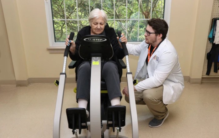 An elderly person receiving guidance from a healthcare professional while using exercise equipment indoors.