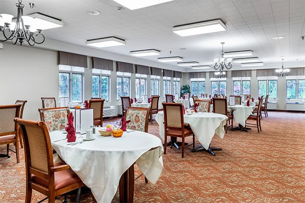 A spacious dining room with elegantly set tables covered with white tablecloths, surrounded by chairs and lit by natural light through large windows.
