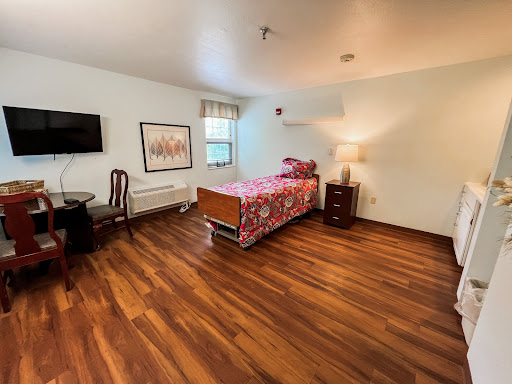 A spacious and well-furnished bedroom featuring a wooden floor, a bed with colorful bedding, a TV mounted on the wall, and a bedside lamp.