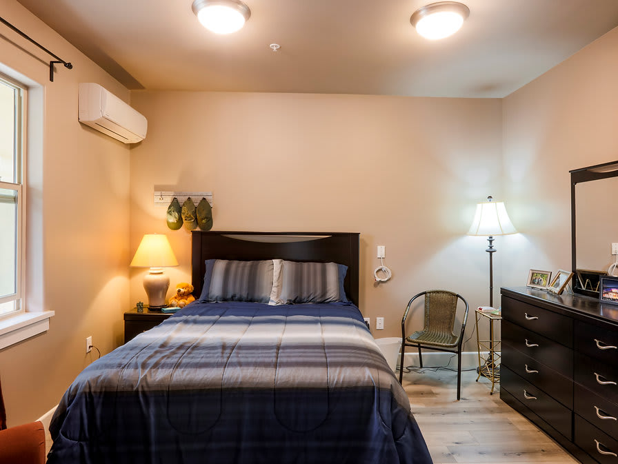 A cozy bedroom with a neatly made bed, flanked by two table lamps, featuring a wall-mounted air conditioner and a simple chair.