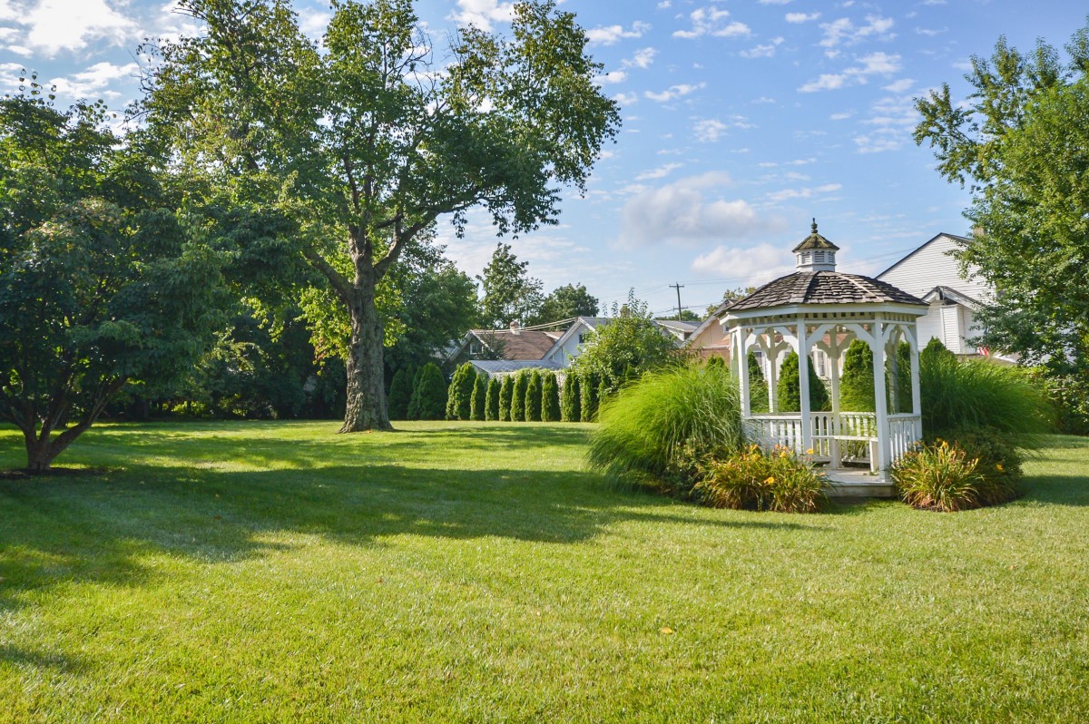 A serene garden scene featuring a white gazebo, lush trees, and a well-maintained lawn under a clear blue sky.
