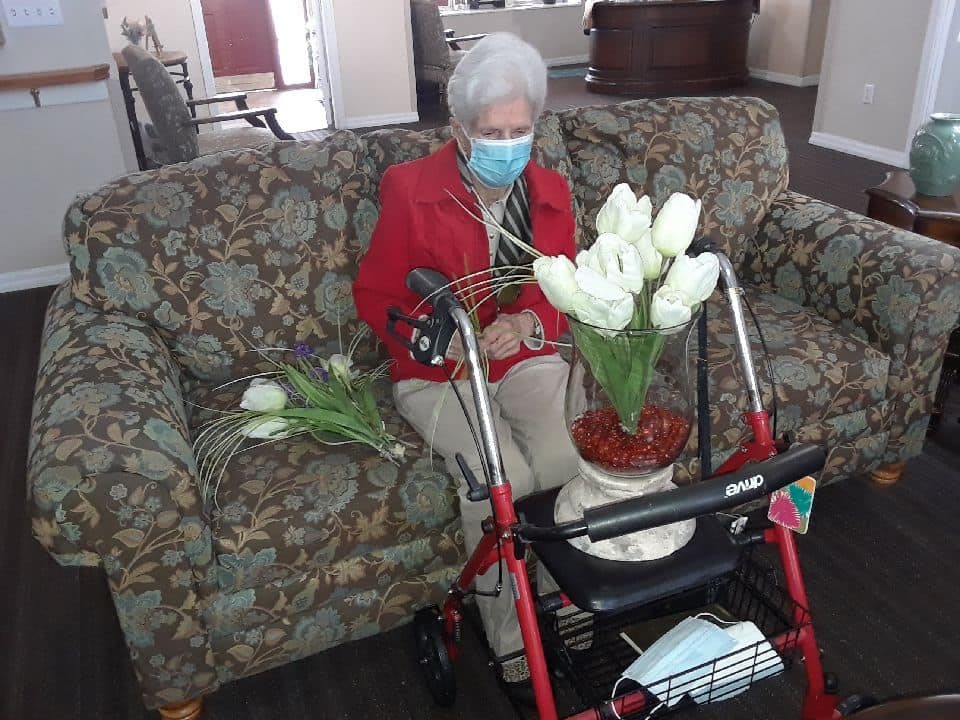 An elderly woman wearing a mask sits on a sofa with flowers and a walker nearby.