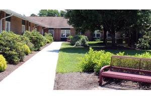 A peaceful garden with a pathway leading to a building, surrounded by greenery and featuring a bench.
