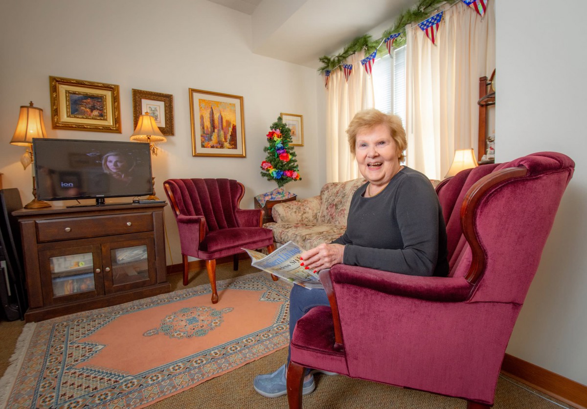 An elderly woman sits comfortably in a cozy living room, holding a book and enjoying a quiet moment.