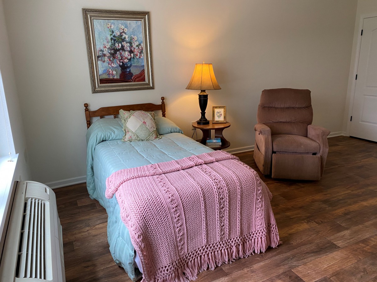 A cozy bedroom featuring a bed with a pink blanket, a recliner chair, a lamp on a side table, and artwork on the wall.