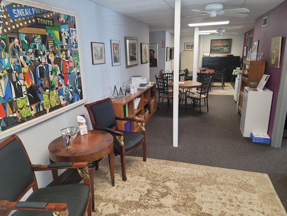 A cozy office interior with a waiting area featuring chairs, tables, artwork on the walls, and a bookshelf.