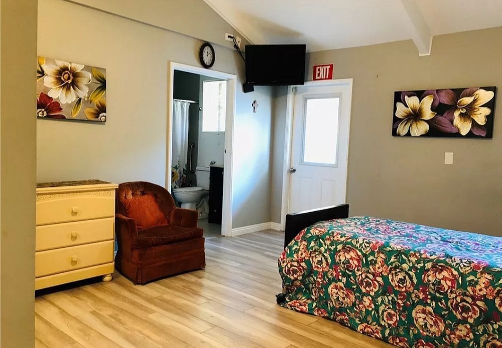 A cozy bedroom with a colorful bedspread, wooden dresser, and an armchair next to a door leading to a bathroom.