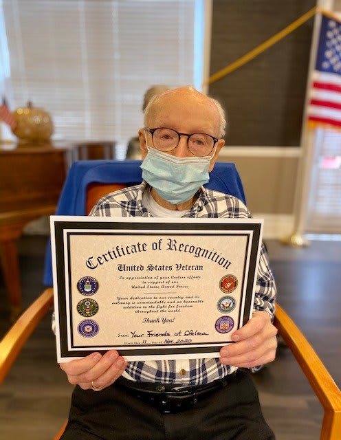 An elderly person holding a certificate of recognition while seated indoors, wearing a mask with an American flag in the background.