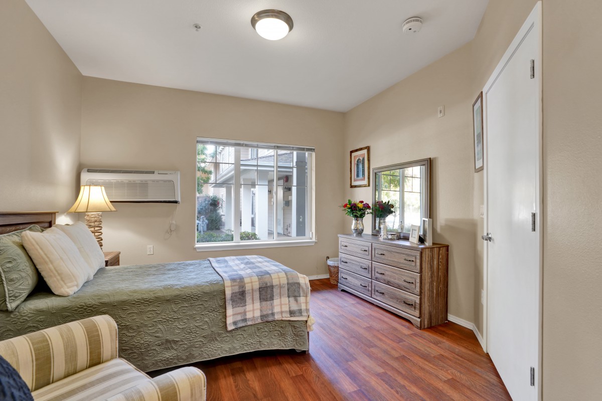 A cozy bedroom with a large window, featuring a bed, dresser with a mirror, and wood flooring.
