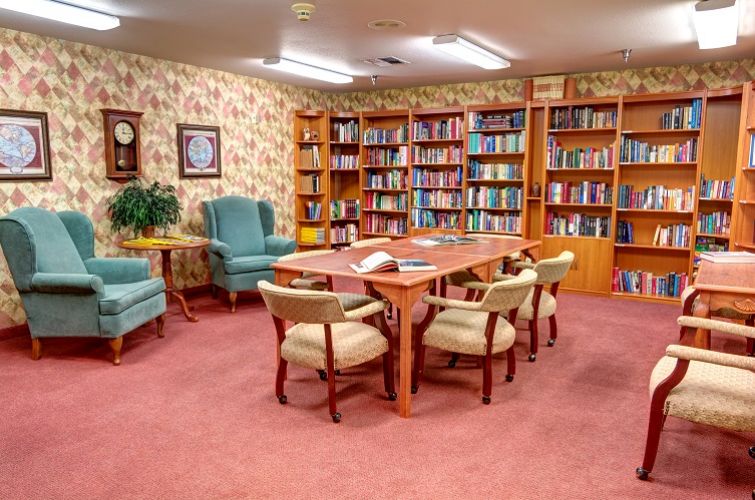 A cozy library room with bookshelves filled with books, a wooden table surrounded by chairs, and two comfortable armchairs.