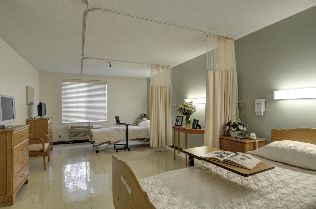 A clean and organized hospital room with two beds, separated by curtains, and a window letting in natural light.