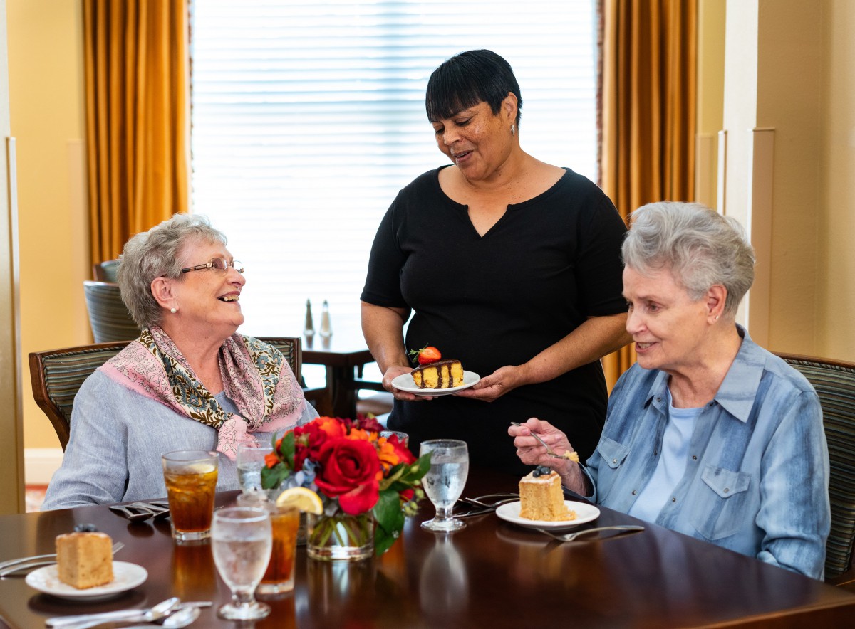 Two elderly women enjoy a meal and laughter as a waitress serves dessert at a restaurant.