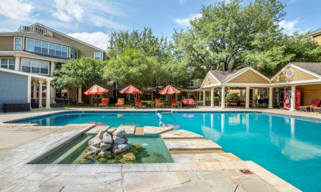 A serene outdoor swimming pool area at an apartment complex with patio seating and lush greenery.