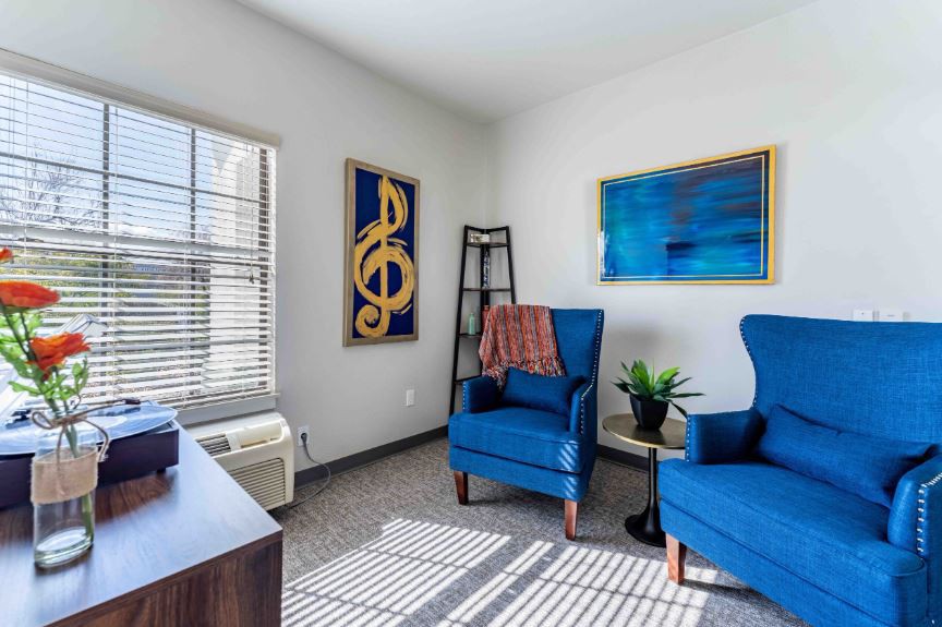 A cozy living room with two blue armchairs, decorated with musical artwork and illuminated by natural light from a window.
