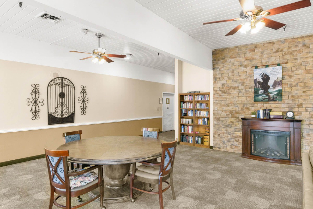 A cozy living space with a dining area, featuring a round table and chairs, a stone fireplace, and bookshelves, complemented by ceiling fans for ventilation.