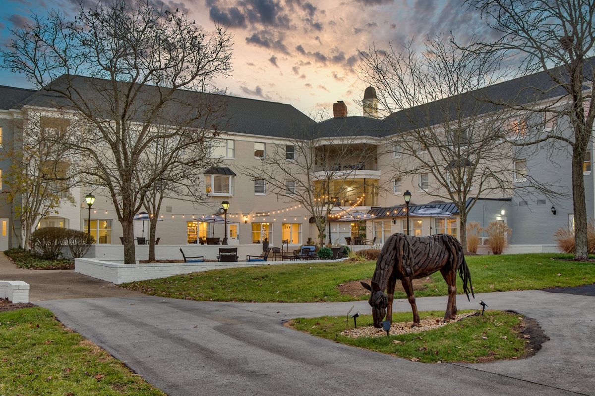 A horse grazes near a building at sunset, surrounded by trees and a pathway.