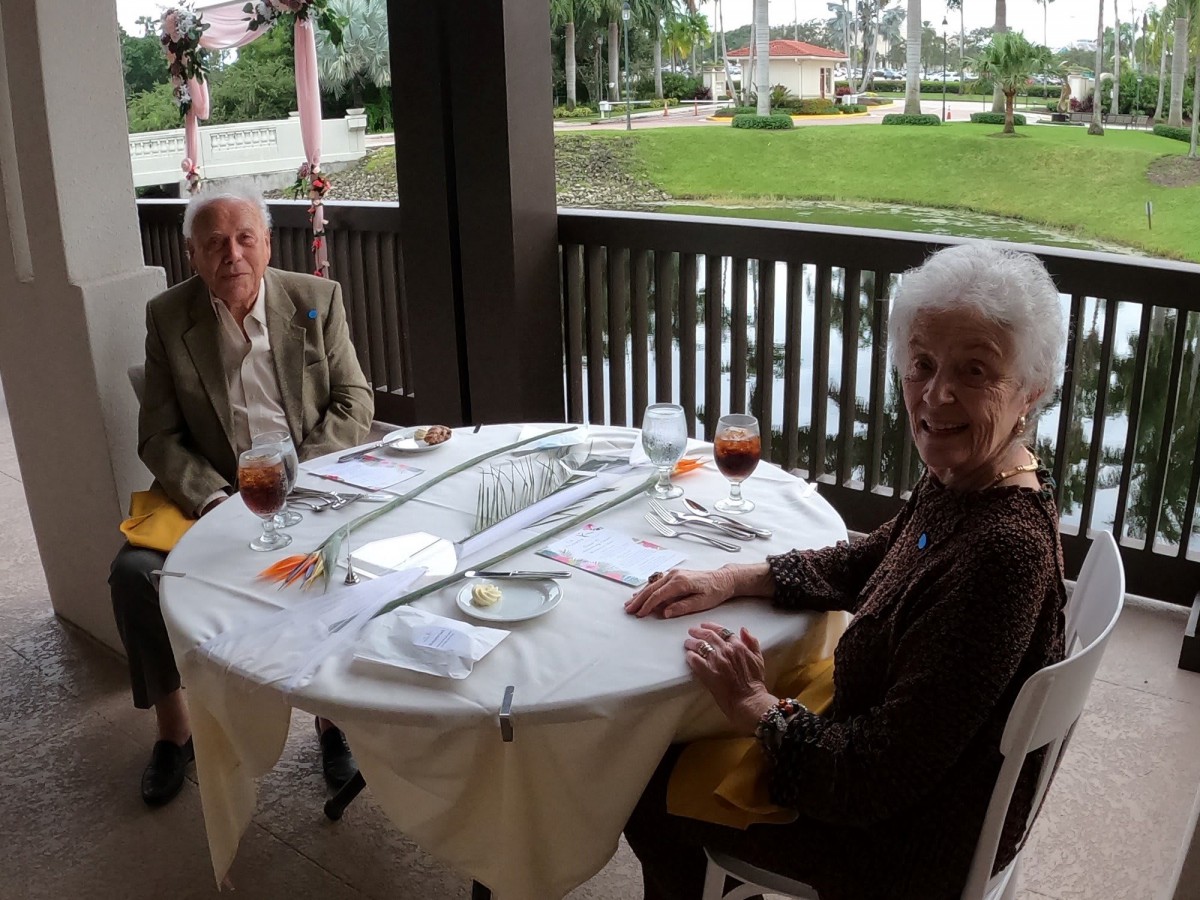 An elderly couple enjoys a meal at an outdoor waterfront restaurant.