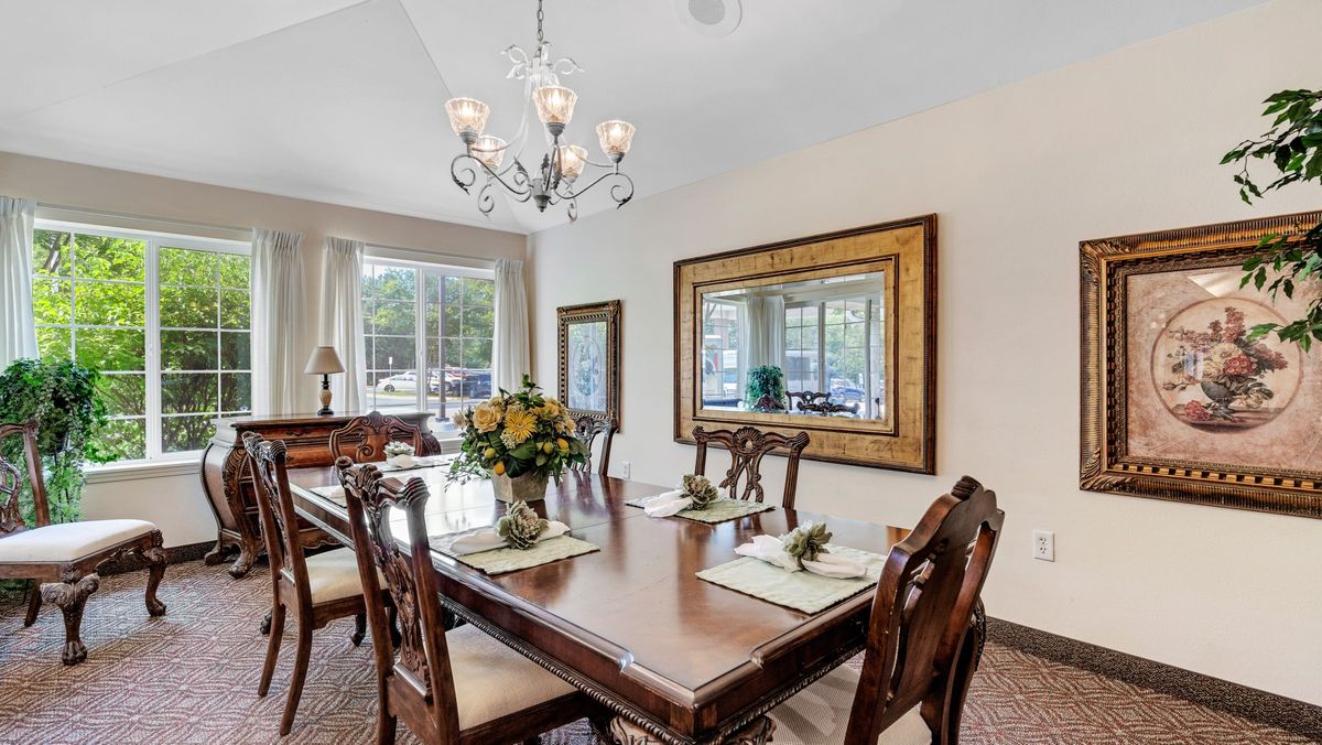 A well-lit dining room featuring a large wooden table with elegant chairs, a chandelier, and decorative elements such as mirrors and flowers.