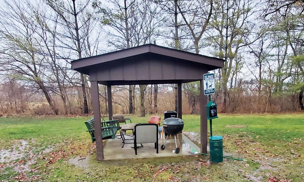 A small outdoor gazebo with chairs and a grill set in a park surrounded by trees.