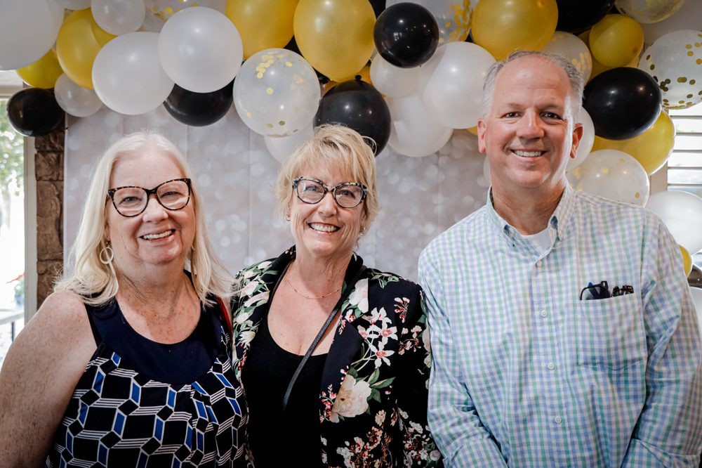 Three people smiling at a celebration with balloons in the background.