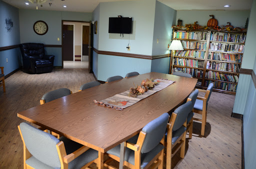 A spacious dining room featuring a large wooden table surrounded by chairs, with a bookshelf and television in the background.