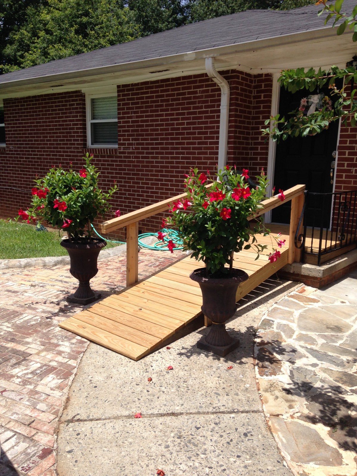 A wooden accessibility ramp leading into a brick house, flanked by two large planters with vibrant red flowers.