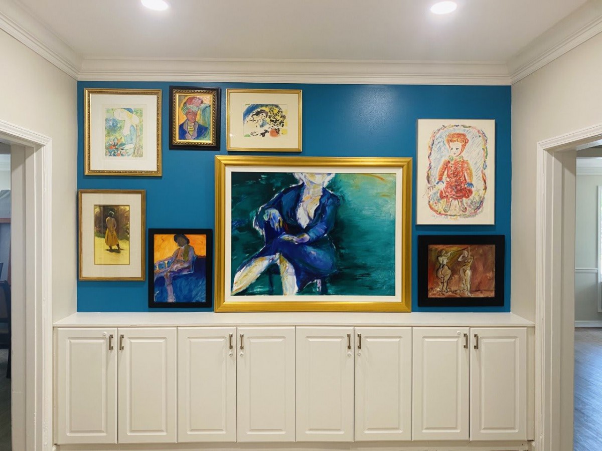 A well-arranged collection of paintings displayed on a blue accent wall above white cabinets, showcasing an eclectic mix of art styles.