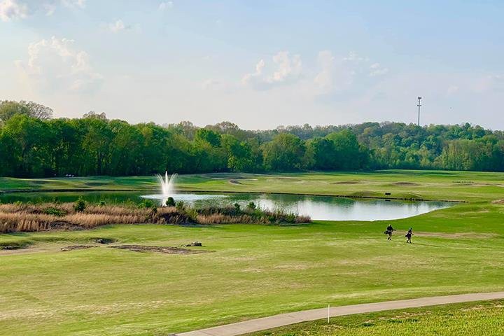 A scenic view of a golf course with a lake and fountain, surrounded by lush greenery.