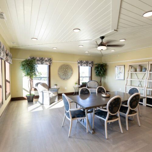 A bright and modern dining room with large windows, a central table surrounded by chairs, potted plants, a bookshelf, and a ceiling fan.