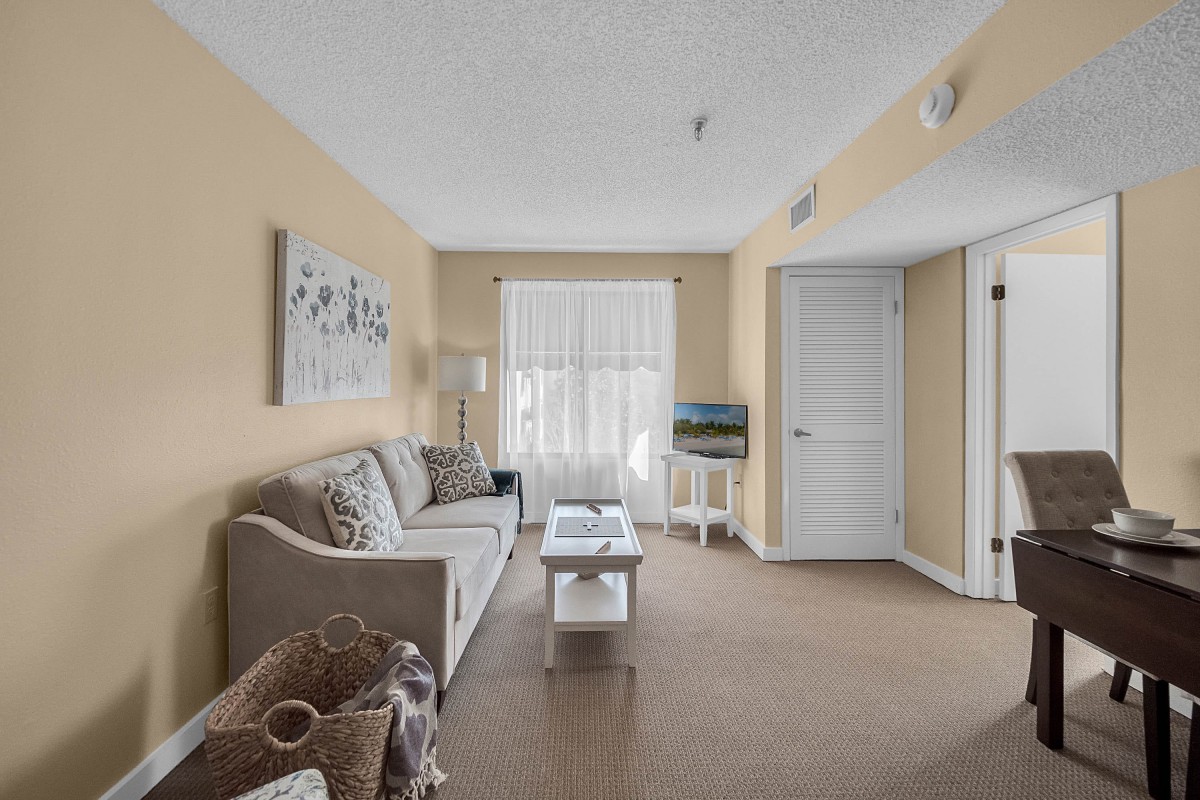 A cozy living room with beige walls, featuring a sofa and coffee table, decorated with wall art and illuminated by natural light through window blinds.