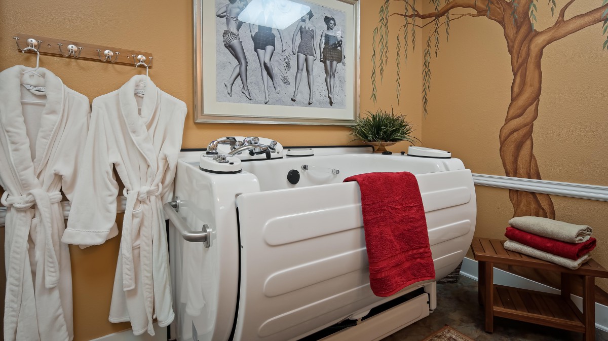 A cozy bathroom featuring a walk-in bathtub with a red towel, robe, and decorative artwork.