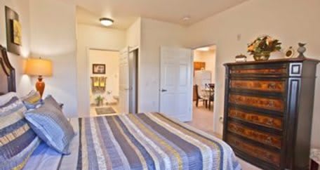 A cozy bedroom with a striped bedspread and wooden dresser, leading to other rooms.